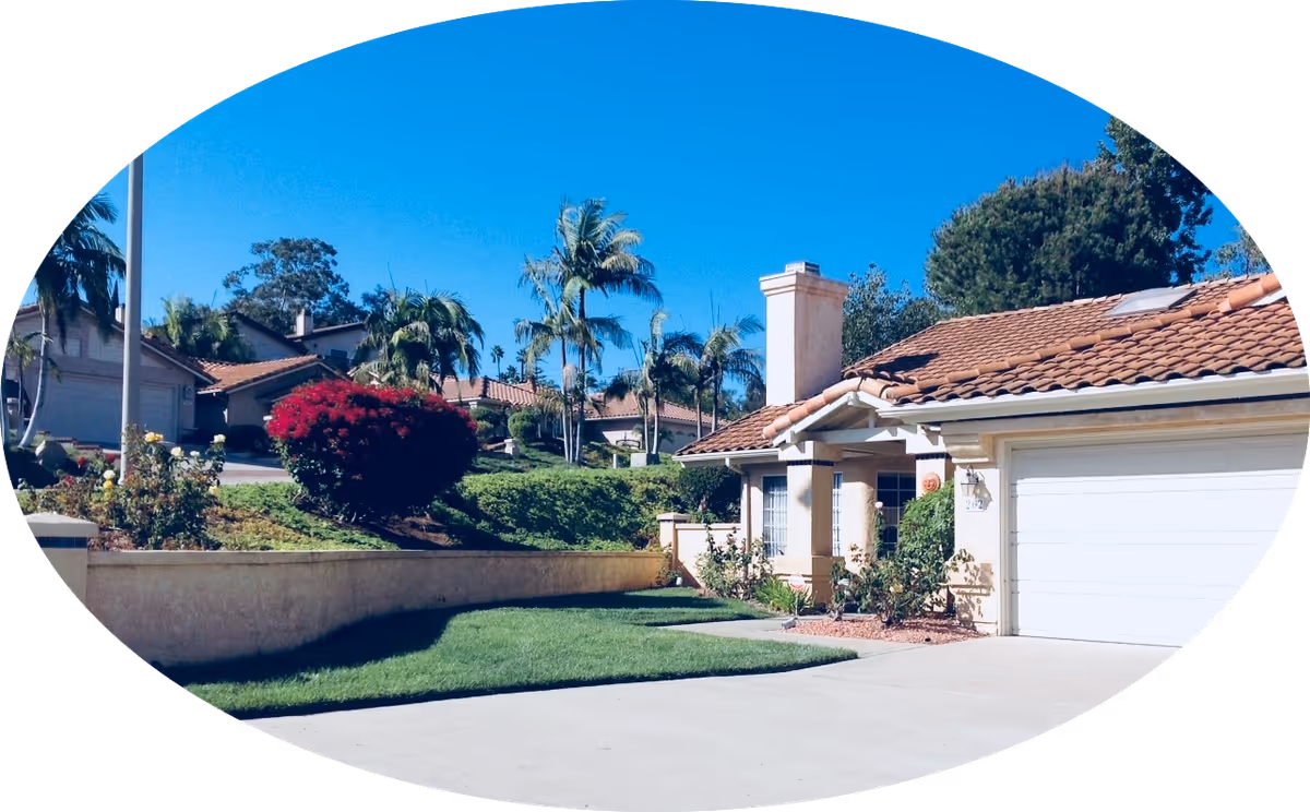 Exterior view of a single-story house with a tiled roof, a white garage door, and a small front porch. The house is surrounded by a well-maintained lawn, bushes, and palm trees under a clear blue sky.