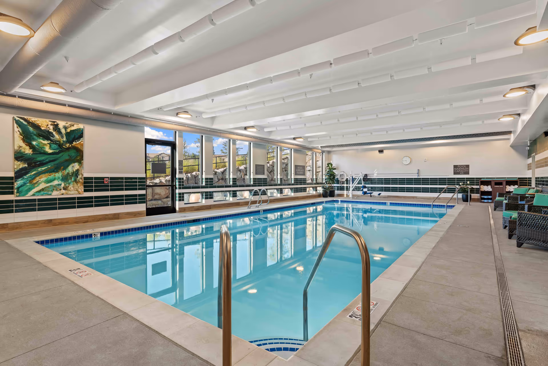 Indoor swimming pool area with clear blue water, metal handrails, lounge chairs with green cushions, large windows letting in natural light, and decorative artwork on the wall.