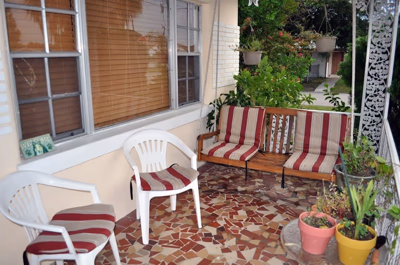 A cozy outdoor porch area with a mosaic tile floor, two white plastic chairs with red and beige striped cushions, a wooden bench with matching striped cushions, several potted plants, and a window with closed blinds. The porch is surrounded by greenery and has a decorative white metal railing.