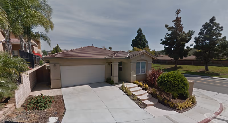 Single-story residential building with a two-car garage, a small front garden with shrubs and trees, and a concrete driveway leading to the street. The house has a tiled roof and beige exterior walls. The surrounding area includes a sidewalk, a street, and some greenery.