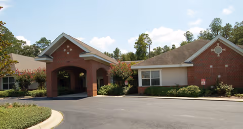 Exterior view of a single-story brick building with a covered entrance and a driveway, surrounded by trees and shrubs under a partly cloudy sky.