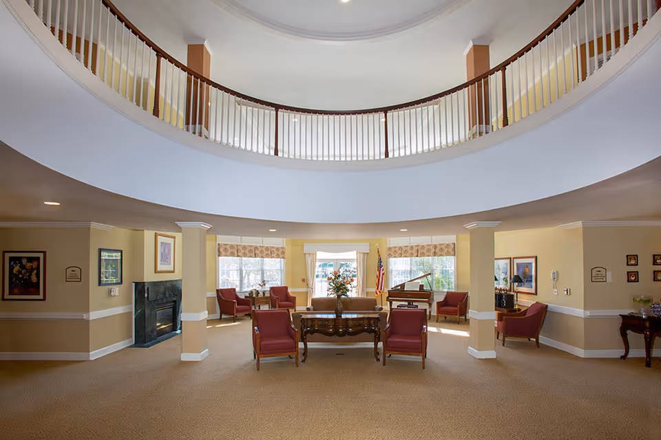 Spacious senior living facility common area with beige walls and carpet, featuring a circular balcony above, multiple red armchairs arranged around wooden tables, a fireplace on the left wall, large windows with patterned valances, and a grand piano near the back right corner.