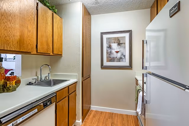 A compact kitchen with wooden cabinets, a white countertop, a stainless steel sink, a dishwasher, a white refrigerator, and a stove. There is a framed abstract artwork on the wall at the end of the kitchen and a wooden floor.