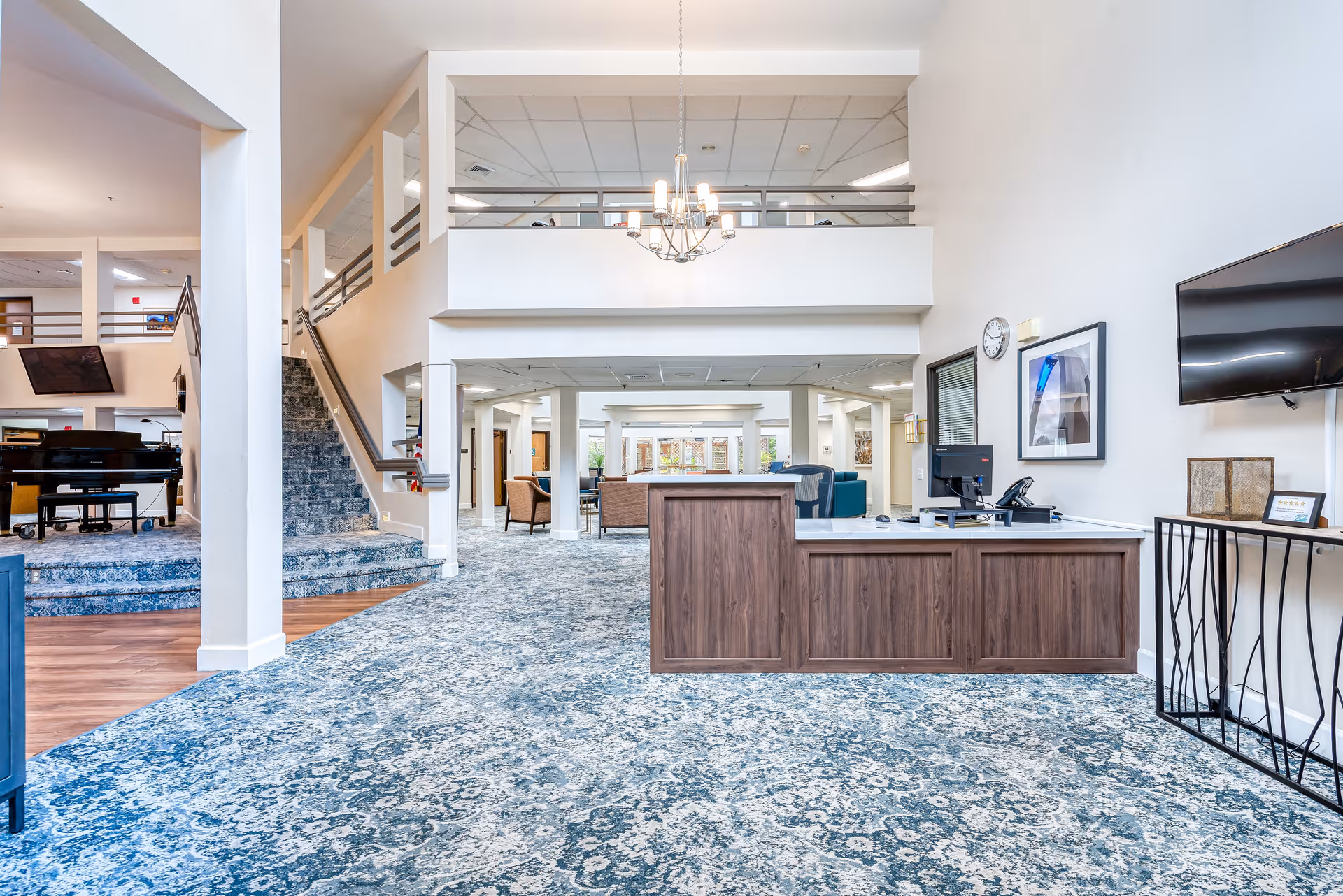 Spacious senior living facility interior with a reception desk in the foreground, blue patterned carpet, a staircase to the left, a grand piano in the background, and seating areas further inside. The space has high ceilings, white walls, and modern lighting fixtures.