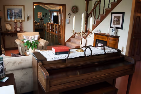 Cozy living room with an upright piano in the foreground, upholstered chairs, a staircase, and decorative furnishings.