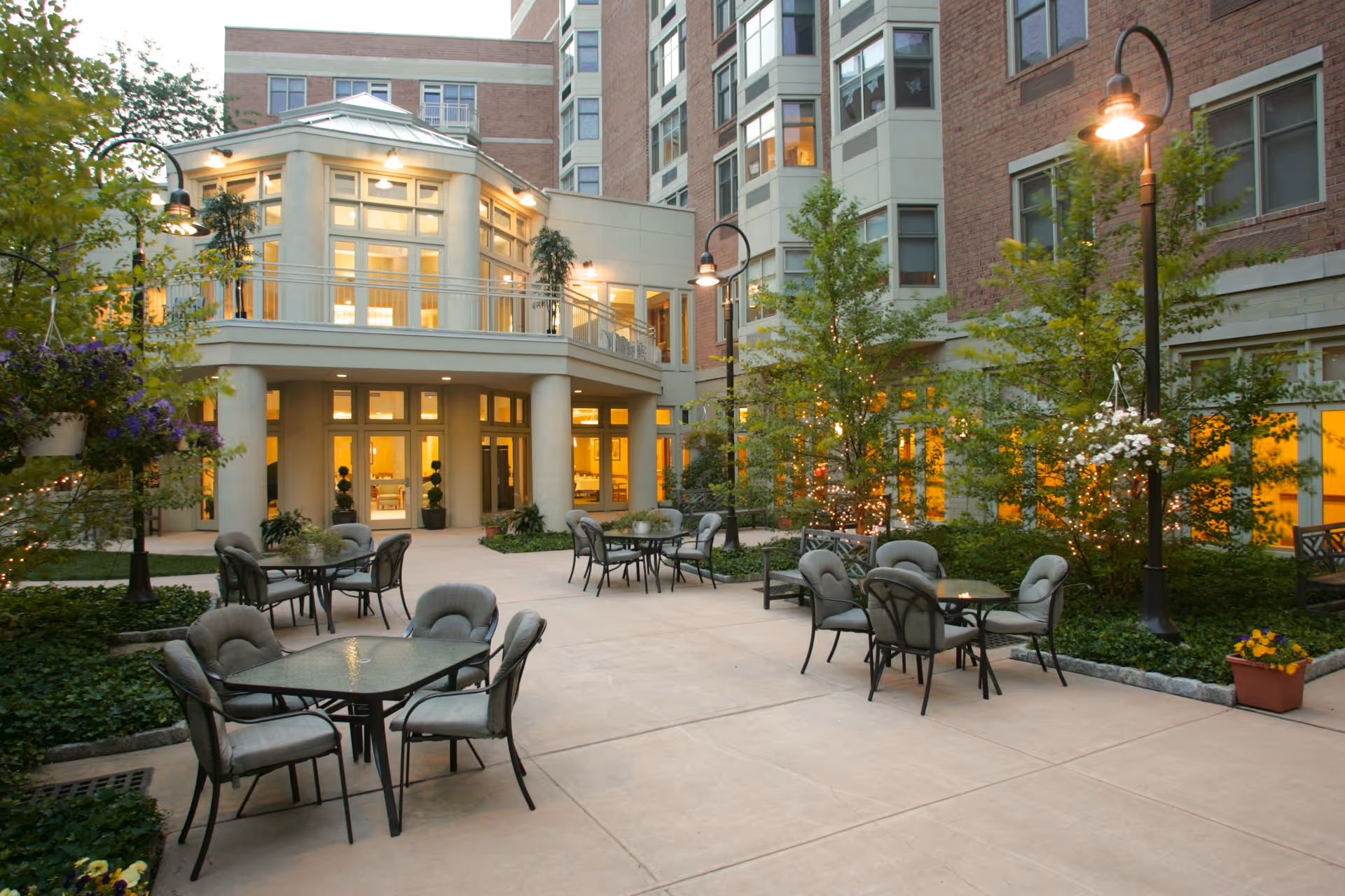 Outdoor courtyard area of a senior living facility with multiple glass-top tables and cushioned chairs arranged on a concrete patio. The courtyard is surrounded by a multi-story building with large windows and warm interior lighting. There are trees and plants with string lights wrapped around them, and several streetlamp-style lights illuminating the space.