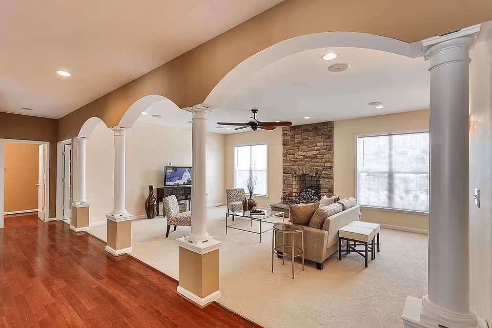 A spacious living room with beige walls and carpet, featuring a stone fireplace, a ceiling fan, a beige sofa with cushions, two patterned armchairs, a glass coffee table, and a TV on a stand. The room is separated from a hallway with wooden flooring by white decorative columns and arches.