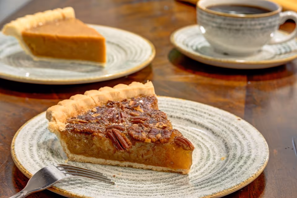 Two slices of pie on ceramic plates on a wooden table, one pecan pie slice in the foreground and one pumpkin pie slice in the background, with a cup of black coffee in a matching ceramic cup and saucer.