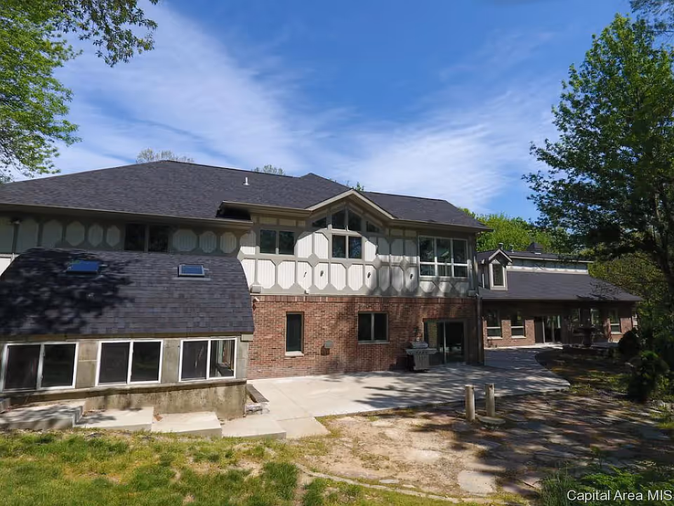 Exterior view of a two-story building with a combination of brick and light-colored paneling under a clear blue sky. The building has multiple windows and a paved area in front, surrounded by trees and greenery.