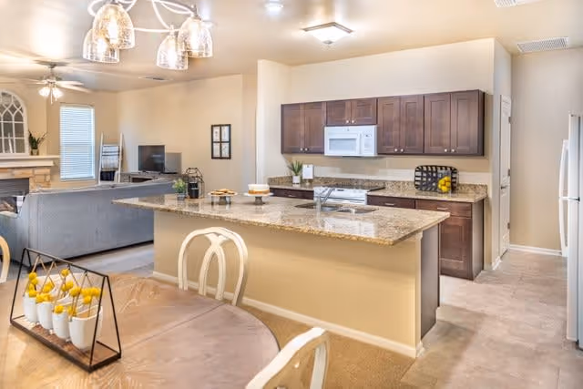 A bright and spacious kitchen and living area in a senior living facility. The kitchen features dark wood cabinets, a granite countertop island with a sink, a white microwave, stove, and refrigerator. In the foreground, there is a wooden dining table with white chairs and a decorative centerpiece with yellow flowers. The living area has a gray sofa, a fireplace, and a TV. The walls are painted beige, and the floor is a mix of carpet and tile.