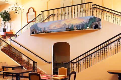 Interior view of a senior living facility showing a staircase with ornate black railings and a mural depicting a mountain landscape with small buildings and tunnels. There are tables and chairs in the foreground, a chandelier hanging from the ceiling, and a large plant in the corner.