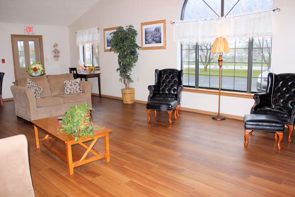 A senior living facility common area with wooden flooring, a beige loveseat with patterned pillows, two black leather armchairs with matching ottomans, a wooden coffee table with a green potted plant, a tall floor lamp, a small table with a lamp and flowers near a window, framed artwork on the walls, and large windows letting in natural light.