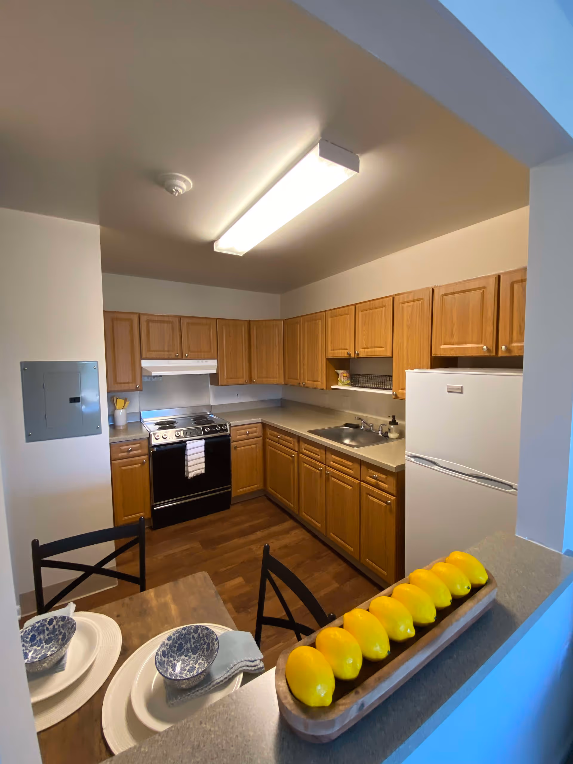 U-shaped kitchen with wood cabinets, a stove and sink, a white refrigerator, and a counter with decorative lemons and a set dining place in the foreground.