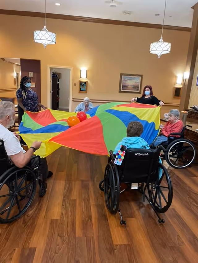Residents and staff in a communal room hold a colorful parachute with balls during a group activity.