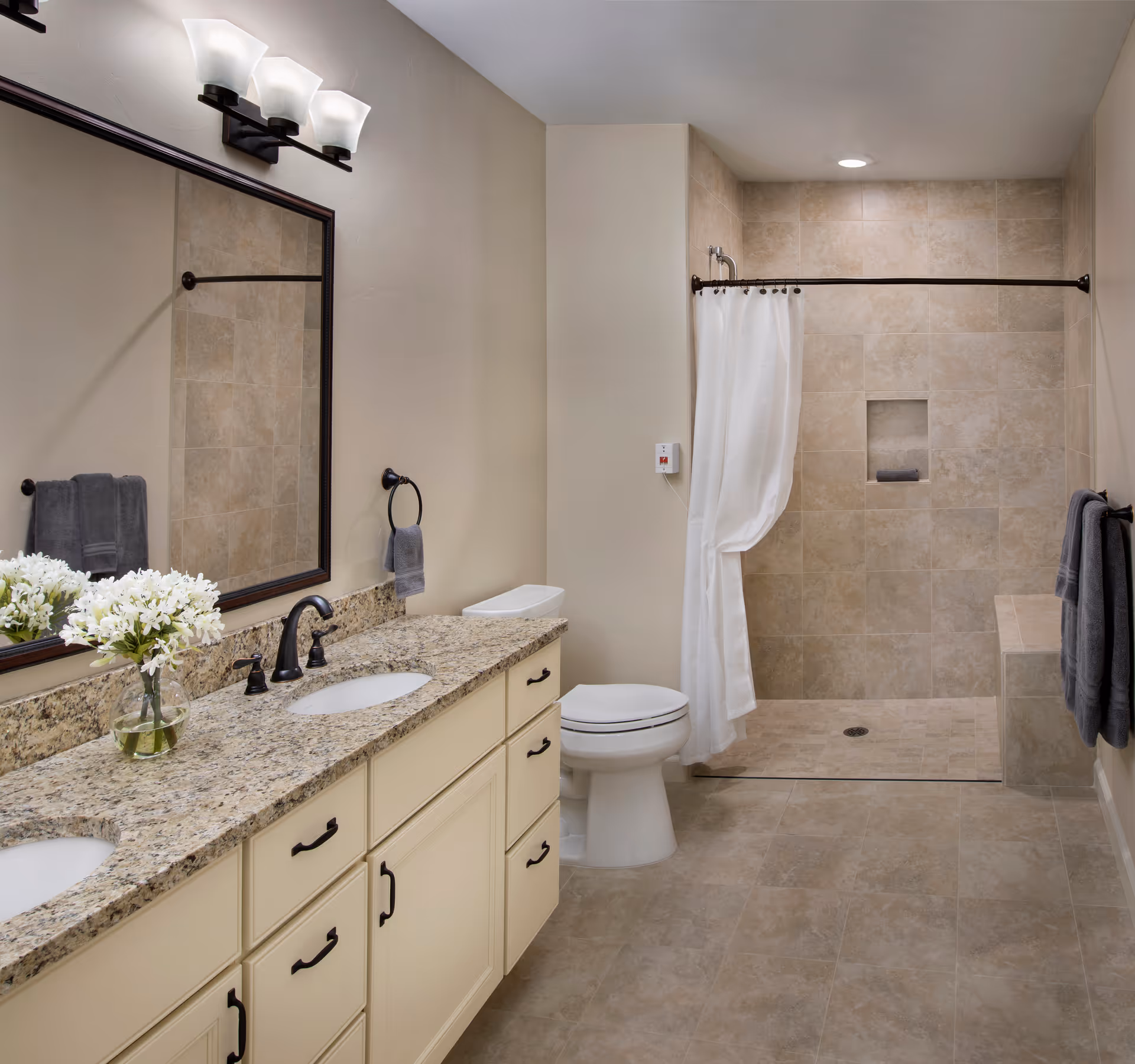 Bright tiled bathroom with a double-sink granite vanity, toilet, and a walk-in shower with a white curtain.