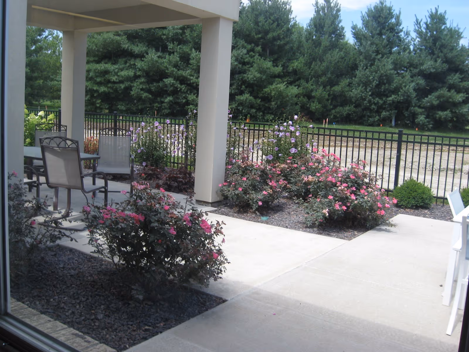 Outdoor patio area with a concrete floor, metal chairs and tables, surrounded by flower beds with pink and purple flowers, and a black metal fence. In the background, there are tall green pine trees under a partly cloudy sky.