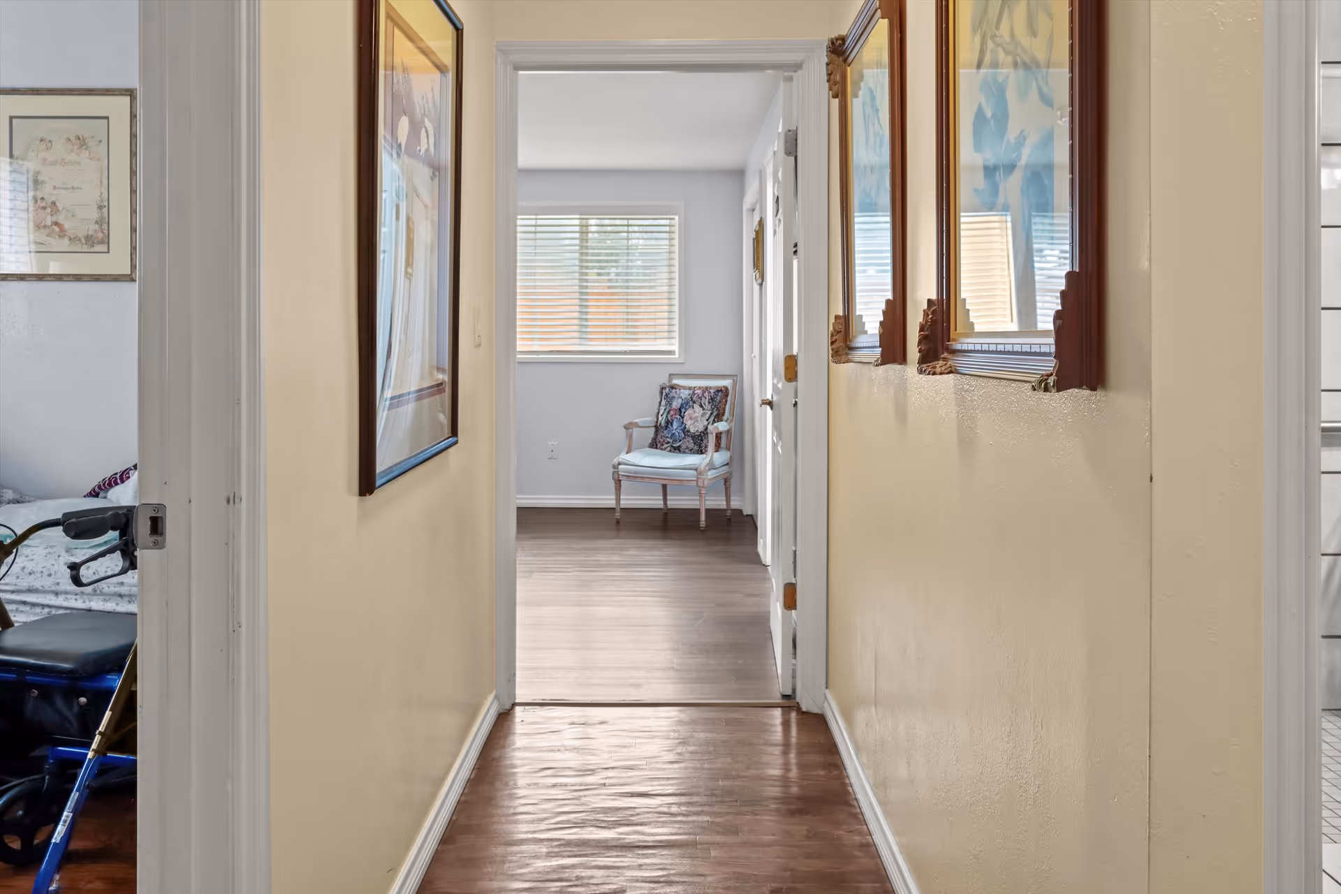 View down a hallway with light yellow walls and wooden floor. There are framed pictures hanging on the right wall and a framed picture on the left wall. At the end of the hallway, there is a room with a window with blinds and a cushioned chair with floral upholstery. To the left, part of a bedroom with a bed and a walker is visible. To the right, a glimpse of a bathroom with white tiles is seen.
