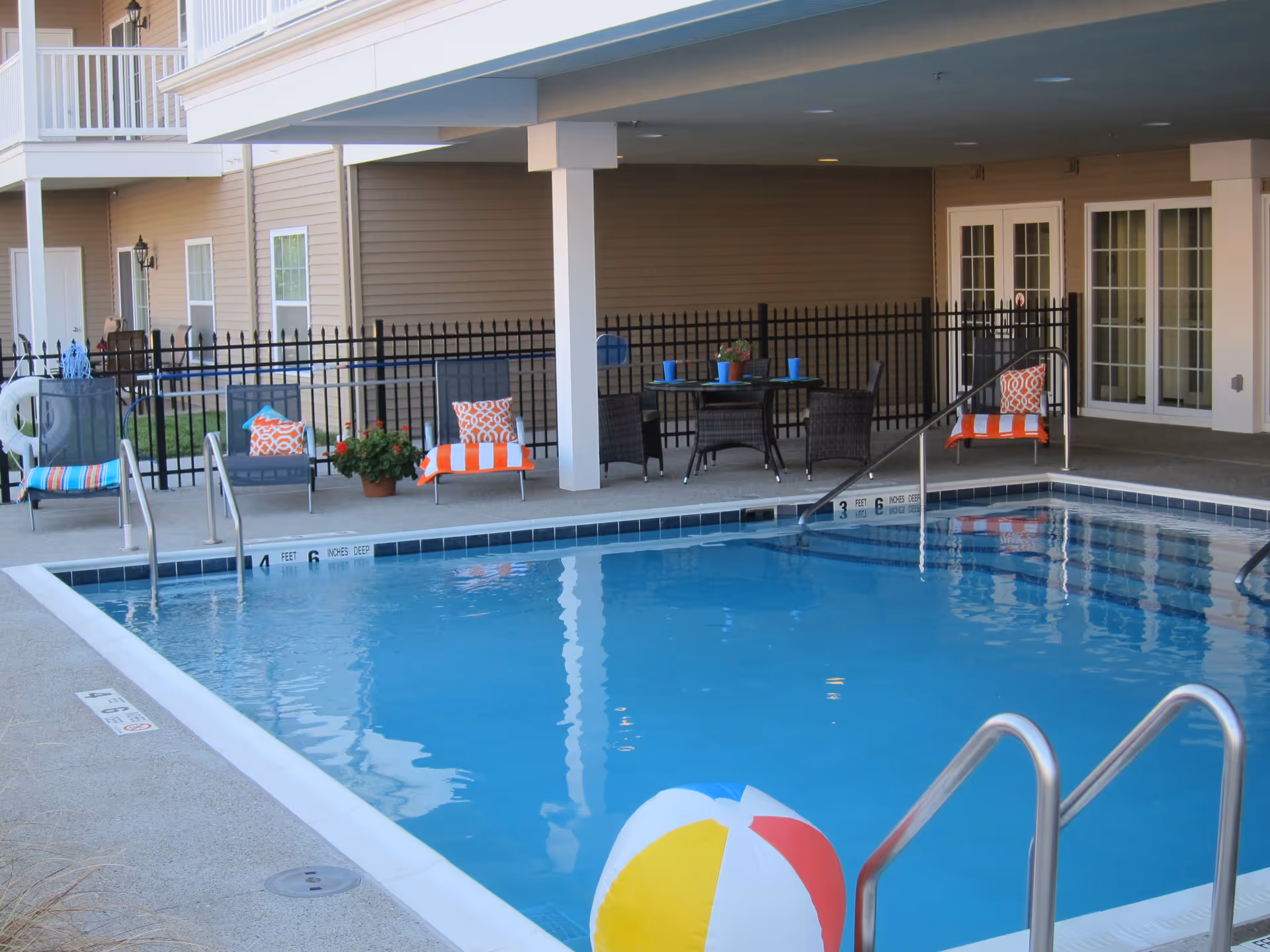Outdoor swimming pool area at Fox Creek Estates with a colorful beach ball floating in the water. Surrounding the pool are lounge chairs with orange and white towels and pillows, a table with chairs, and a black metal fence. The pool area is partially covered by the building structure.