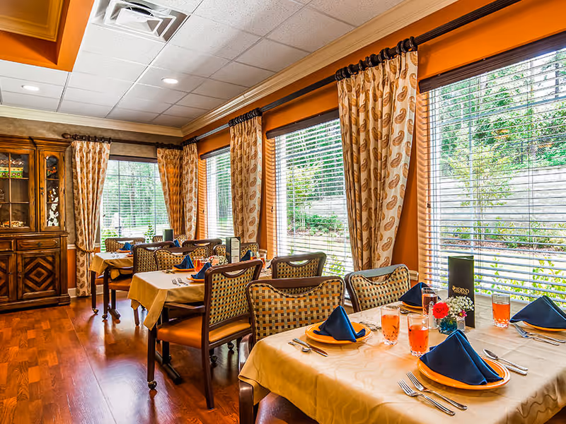A bright dining room with several tables covered in beige tablecloths, each set with orange plates, blue folded napkins, silverware, and glasses filled with a pink beverage. The room has large windows with patterned curtains allowing natural light to enter, and a wooden cabinet is visible in the corner.