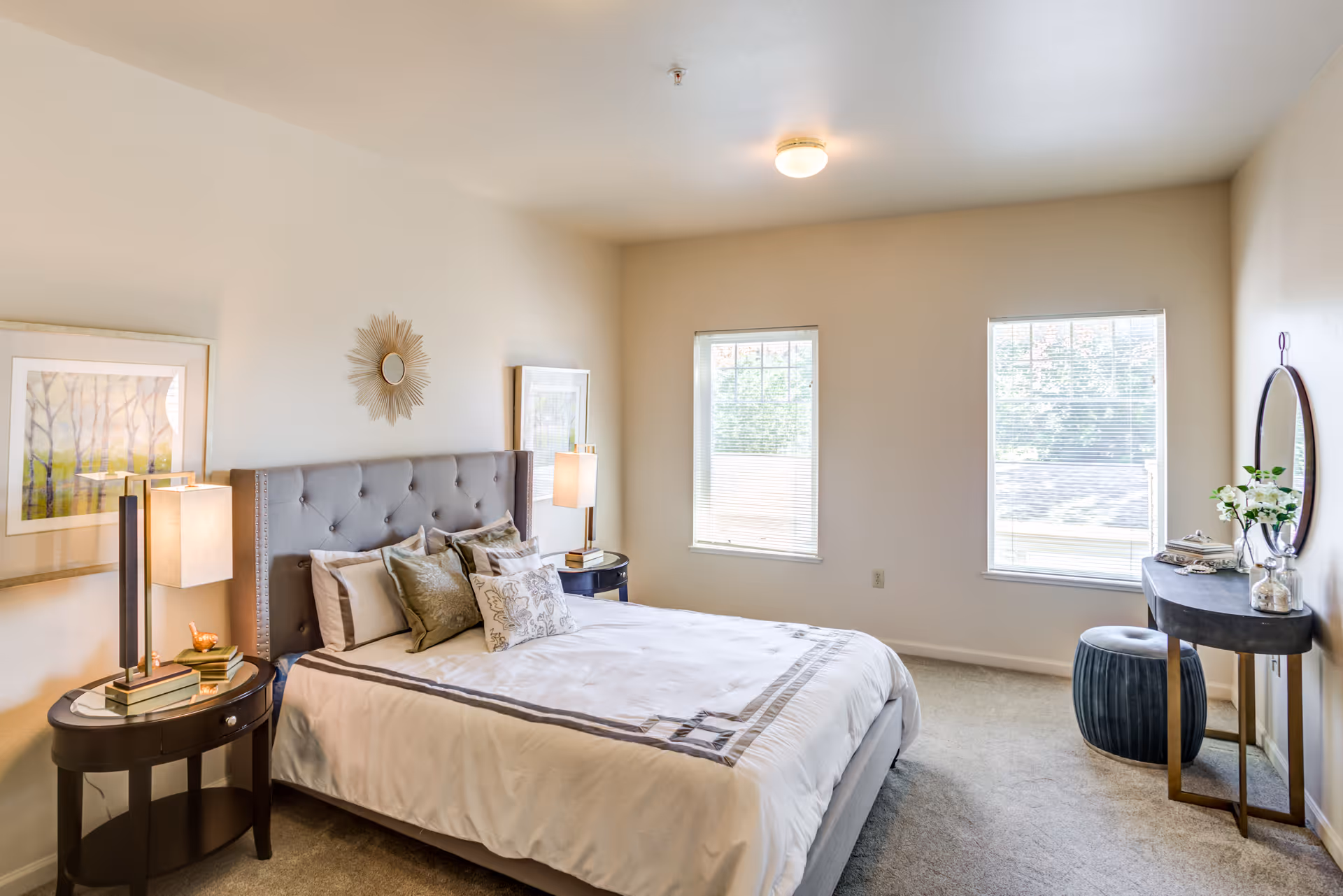 A bright bedroom with a large bed featuring a tufted headboard and multiple pillows. There are two windows letting in natural light, two bedside tables with lamps, framed artwork on the walls, and a small vanity table with a round mirror and a cushioned stool.