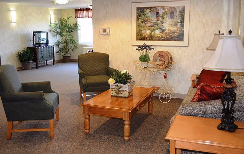 A cozy living room area with two green armchairs, a patterned sofa with red pillows, a wooden coffee table with a floral centerpiece, a side table with a lamp, a small TV on a stand, potted plants, and a framed painting on the wall.