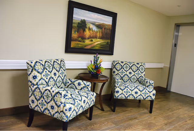 Two patterned armchairs flanking a small round table with a flower arrangement beneath a landscape painting in a hallway seating area.