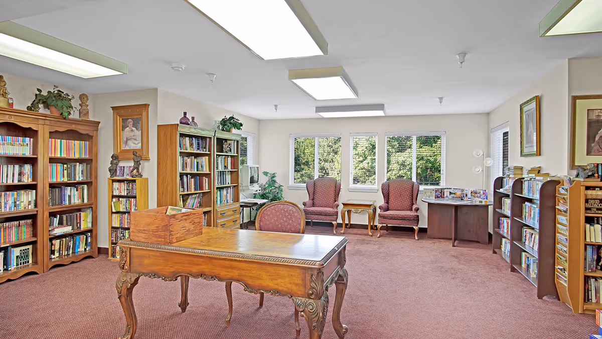 A well-lit library room with wooden bookshelves filled with books, a wooden table with carved legs, a chair, two upholstered armchairs near windows, and framed artwork on the walls. The room has carpeted flooring and large windows showing greenery outside.