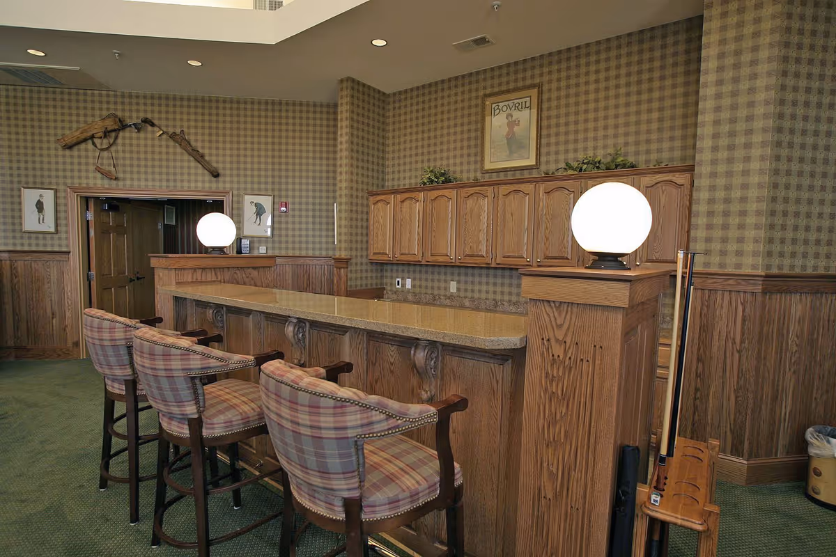 Interior view of a room with a wooden bar counter and four plaid upholstered bar stools. The walls are covered with checkered wallpaper and wood paneling. Two round globe lamps sit on the bar counter. There are framed pictures and a mounted antique rifle on the wall. A pool cue rack with cues is visible on the right side.