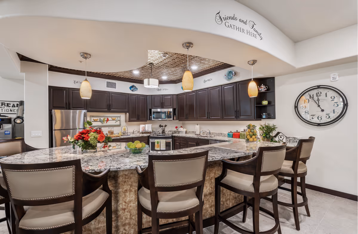 A modern kitchen with dark cabinets, a granite countertop, and bar seating. The space features decorative elements like a large clock, colorful accents, and a bowl of fruit on the counter.