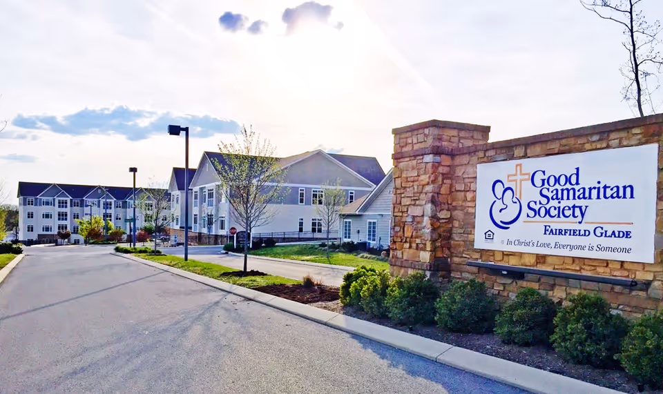 Exterior view of Good Samaritan Society - Fairfield Glade facility with a stone sign displaying the facility name and slogan. The building is a large, multi-story structure with white siding and multiple windows, surrounded by landscaped greenery and a paved driveway.