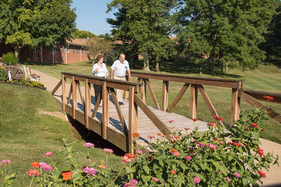 Two older adults walking across a wooden footbridge on landscaped grounds with flowering plants and trees.