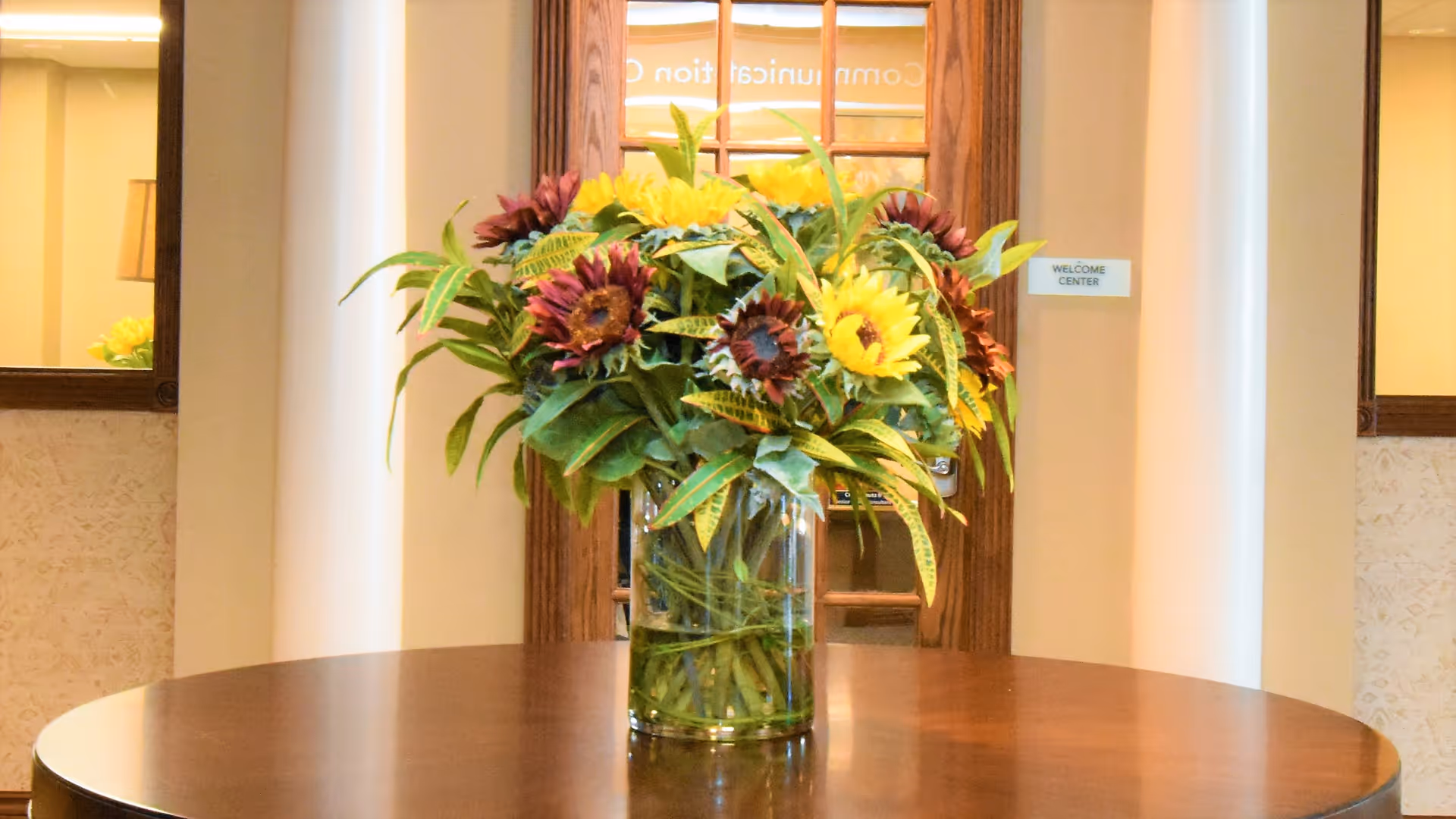 A glass vase of sunflowers and greenery sits on a round wooden table in an interior lobby area with a welcome center door behind it.