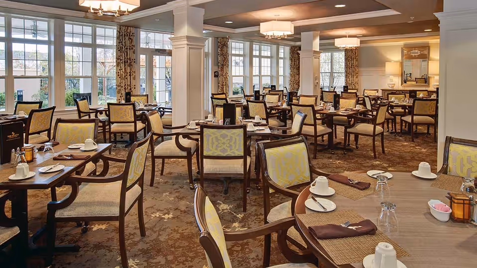 Spacious dining room with multiple set tables and upholstered chairs, large windows, and warm lighting.