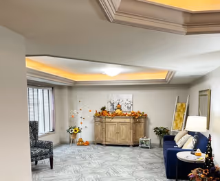 A cozy living room area with a blue sofa, a side table with a lamp, and a wooden cabinet decorated with autumn-themed items including pumpkins and flowers. There is a patterned armchair near a window with blinds, and a framed picture on the wall. The ceiling has recessed lighting with a tray ceiling design.