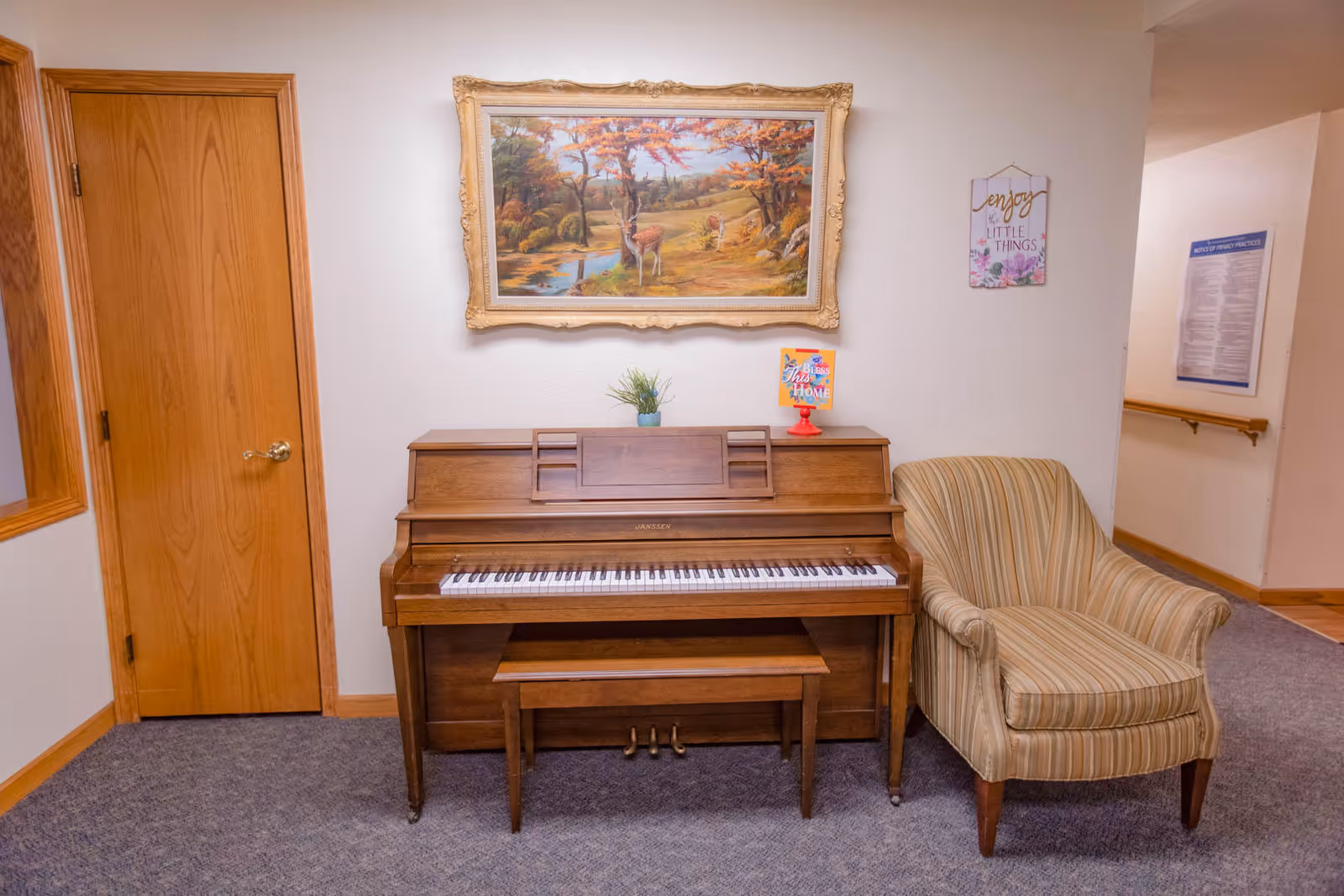 A wooden upright piano with a bench sits against a wall under a framed landscape painting, next to a striped armchair and a closed wooden door.