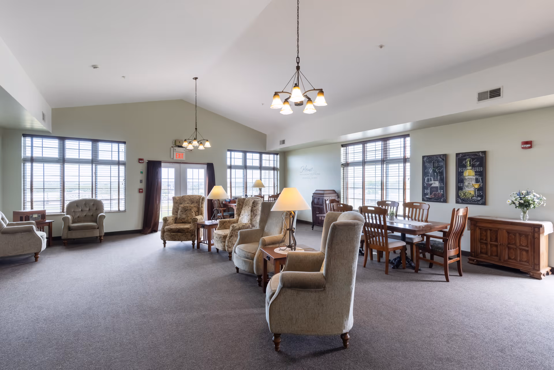 A spacious, well-lit common area in a senior living facility featuring multiple upholstered armchairs arranged around small tables with lamps. There is a wooden dining table with chairs near large windows with blinds, and decorative wall art above a wooden sideboard. The room has high ceilings with hanging light fixtures and carpeted floors.