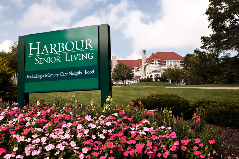 A green sign for Harbour Senior Living, including a Memory Care Neighborhood, is displayed in front of a senior living facility building with a red roof. The foreground features colorful pink and white flowers, and there are trees and a grassy area surrounding the building under a partly cloudy sky.