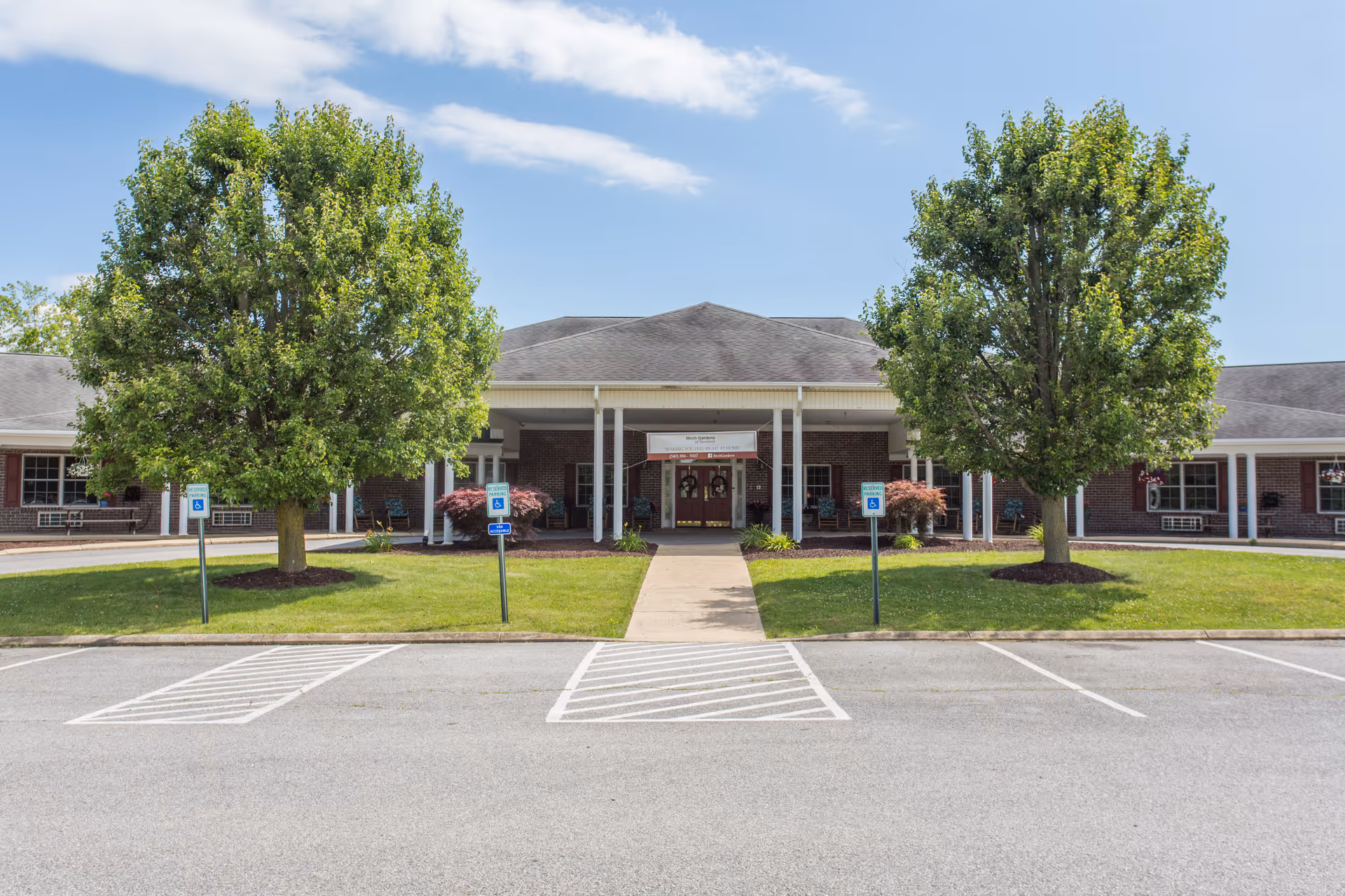 Front entrance of a single-story brick assisted living building with a covered walkway, two large trees, and accessible parking spaces.