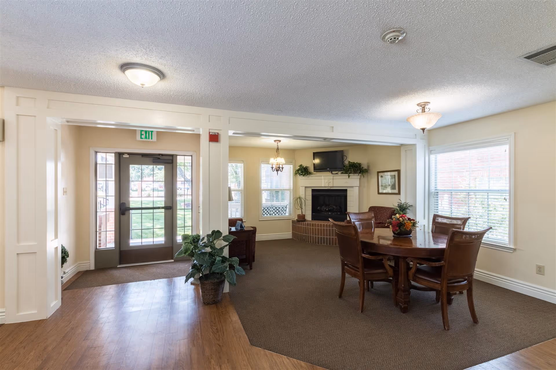 Interior view of a senior living facility common area with a wooden table and four chairs, a fireplace with a TV mounted above it, windows with blinds, and a glass door exit leading outside. The room has beige walls, carpeted and wooden flooring, and ceiling lights.