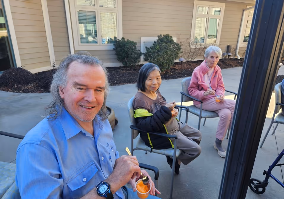 Three elderly people sitting outside on chairs near a building, enjoying cups of orange sherbet or ice cream. The man in the foreground is smiling at the camera, while the two women behind him are also eating and sitting comfortably.