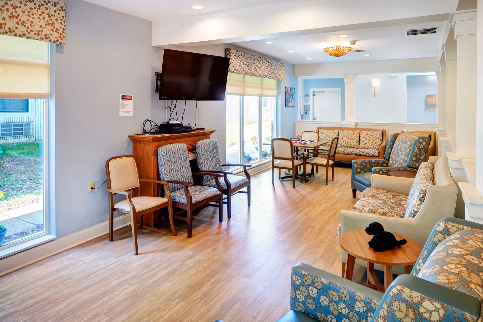 A bright and cozy common area in a senior living facility featuring multiple patterned armchairs, a wooden table with chairs, a wall-mounted flat-screen TV above a wooden cabinet, large windows with floral valances, and a small black stuffed dog on a round wooden side table.