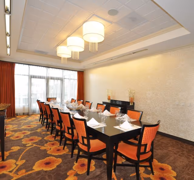 A dining room with a long black rectangular table set for a meal, surrounded by orange cushioned chairs. The room has large windows with sheer white curtains and orange drapes, a floral patterned carpet, and three modern ceiling lights.