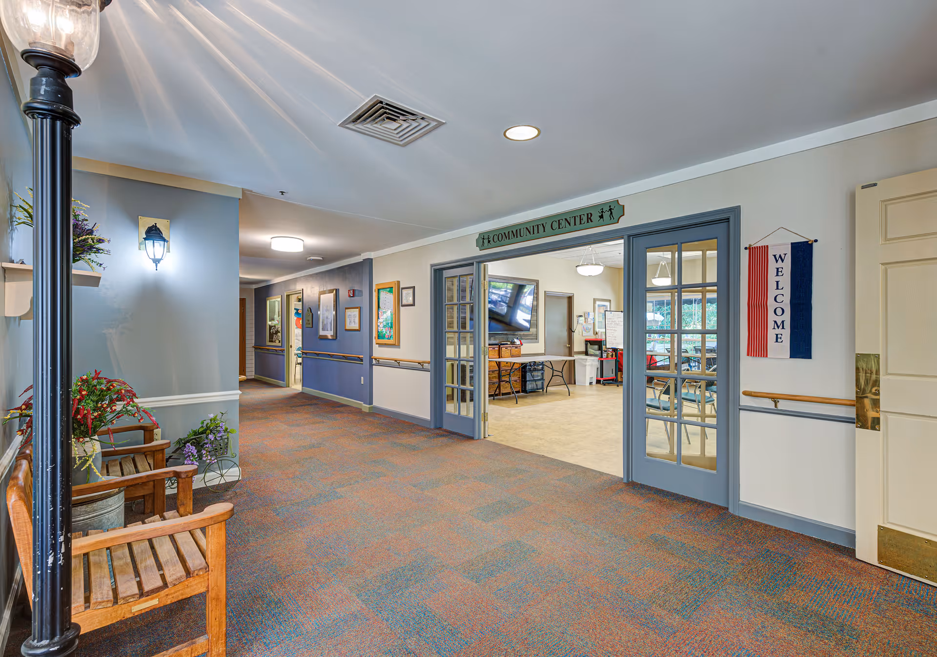 Interior hallway of a senior living facility with carpeted floor and blue-gray walls. On the left, there are wooden benches with potted plants and a decorative street lamp. Ahead, double glass doors lead into a community center room with tables, chairs, and a large TV mounted on the wall. A green sign above the doors reads 'Community Center' and a red, white, and blue welcome banner hangs on the right wall.