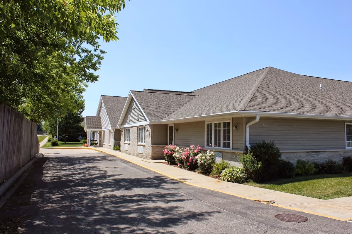 Exterior view of a single-story building with a sloped roof, beige siding, and brick accents. There are windows and a door along the side, with a sidewalk and landscaped bushes and flowers in front. A tree and wooden fence are visible on the left side, and the sky is clear and blue.