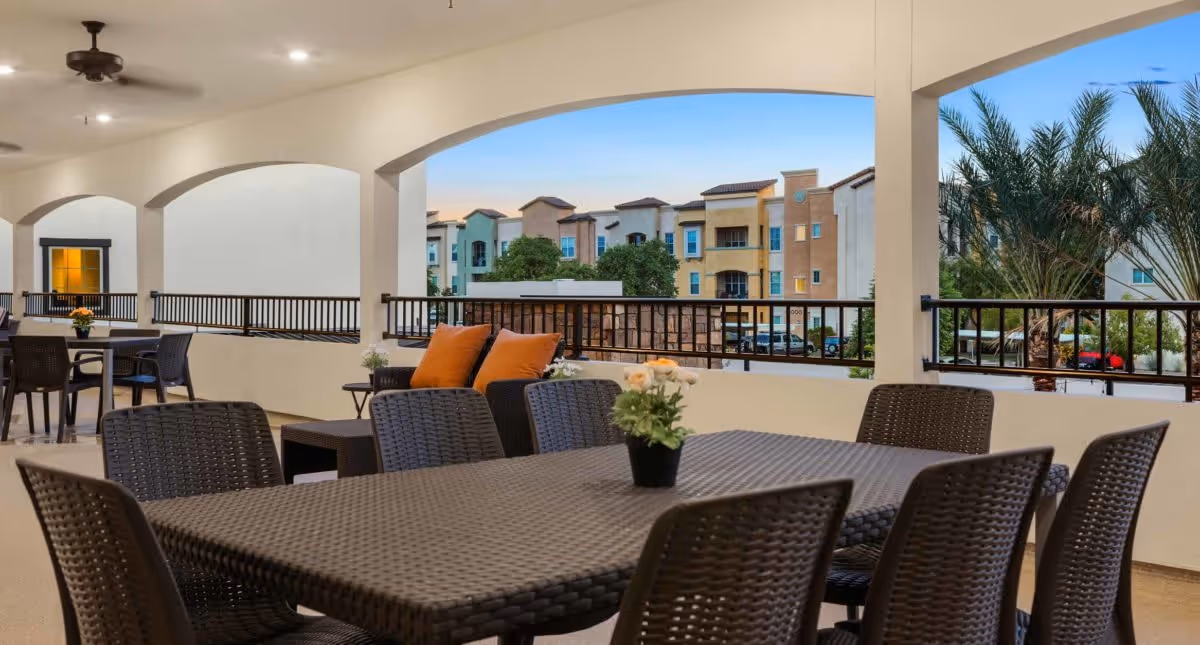 Covered outdoor patio area with multiple dark wicker tables and chairs, some tables decorated with small flower pots. The patio has arched openings with black railings, overlooking a view of colorful apartment buildings and palm trees under a clear sky.