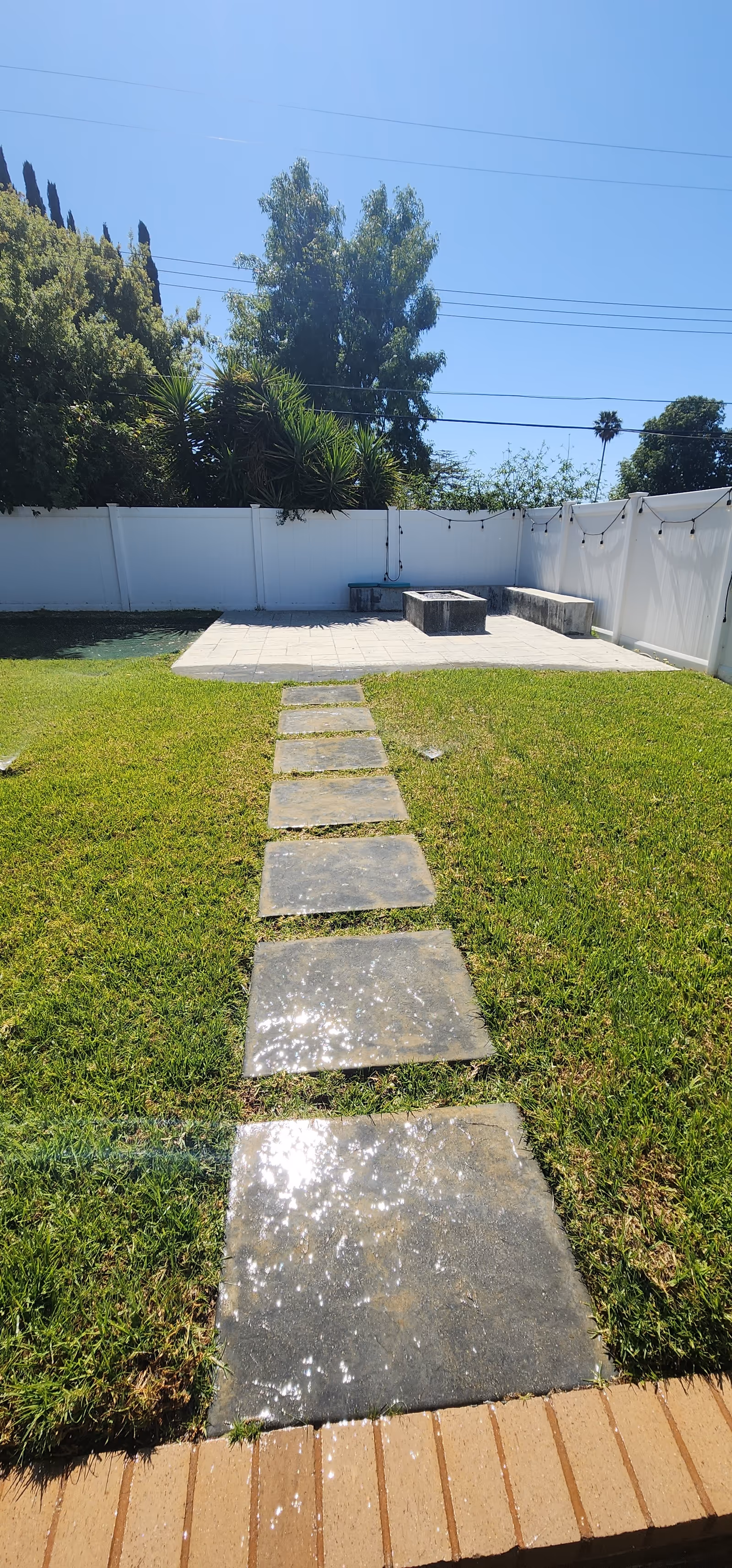 A backyard area with a stone pathway leading across a green lawn to a small paved patio. The patio has built-in concrete benches and a fire pit in the center. The area is enclosed by a white fence, with trees and shrubs visible beyond the fence under a clear blue sky.