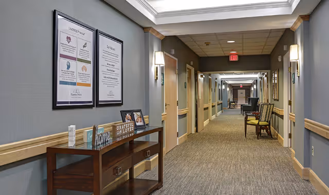 Long carpeted interior hallway in a senior living facility with framed signs, a console table and chairs along the walls.