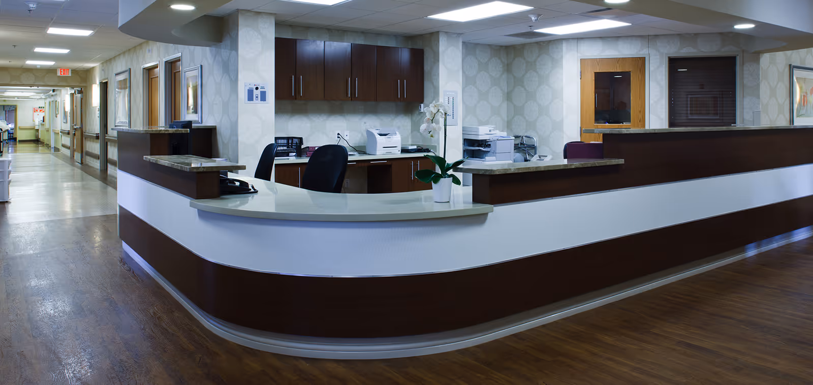 Reception desk area inside a senior living facility with a curved counter, office chairs, cabinets, a printer, and a potted orchid plant. The hallway extends to the left with doors and handrails along the walls.