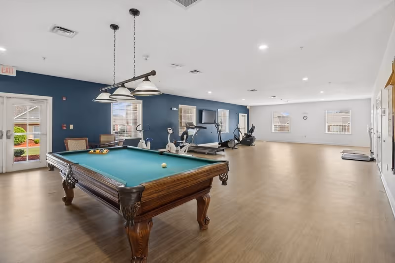 A spacious recreational room with a pool table in the foreground and exercise equipment including treadmills and stationary bikes along the back wall. The room has wooden flooring, white and blue walls, multiple windows letting in natural light, and ceiling lights. There are chairs near the windows and a door leading outside.