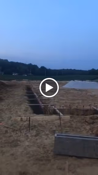 Construction site with dug foundation trenches, rebar, and a concrete block in a sandy field at dusk.